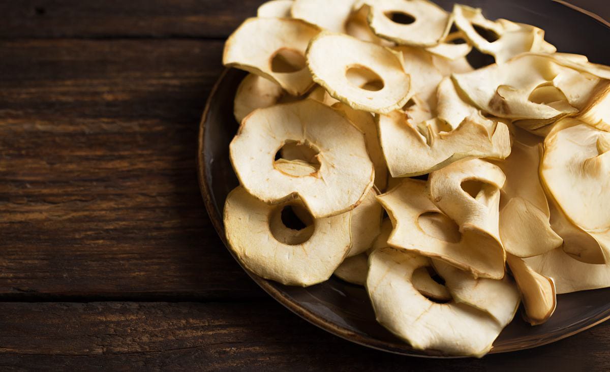 A plate of evenly sliced, golden dried apple rings on a rustic wooden table, showing the results of dehydrating apples without browning.