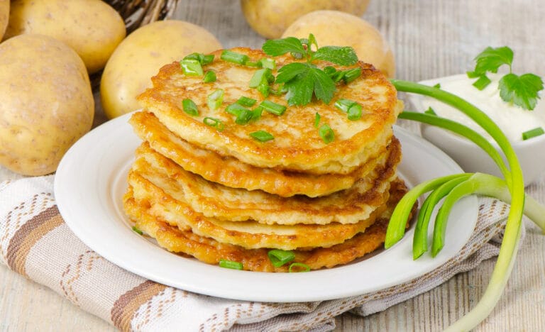 Golden brown leftover mashed potato pancakes stacked on a white plate, garnished with fresh parsley and chopped green onions.