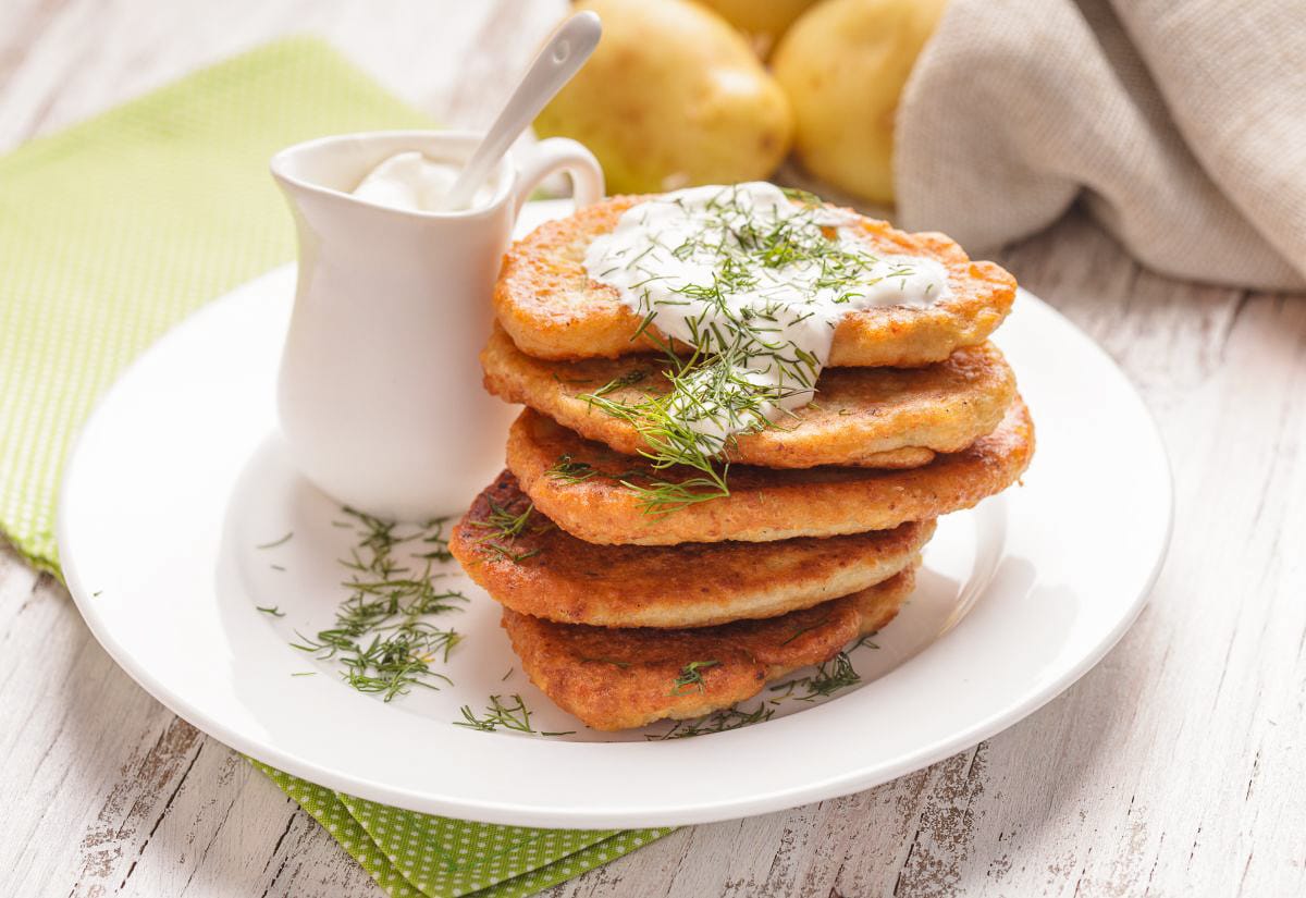 Stack of crispy leftover mashed potato pancakes topped with sour cream and fresh dill on a white plate, served with a small pitcher of sour cream.
