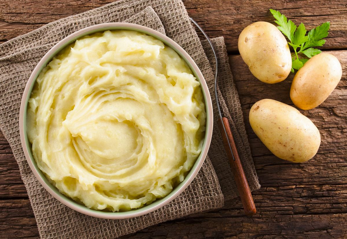 Bowl of creamy leftover mashed potatoes on a rustic wooden table, surrounded by fresh potatoes and a serving spoon.