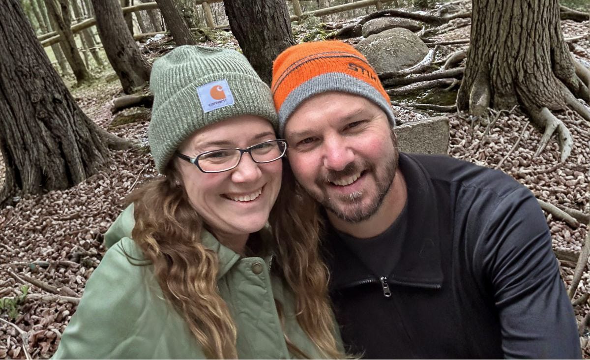 Jessica Knowles and her partner Michael smiling together in a wooded area, wearing knit hats and outdoor clothes on a cool day in Maine.