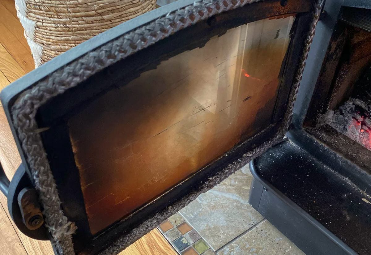 Wood stove door open to show soot-darkened glass, showing what happens when a stove burns inefficiently or needs cleaning.
