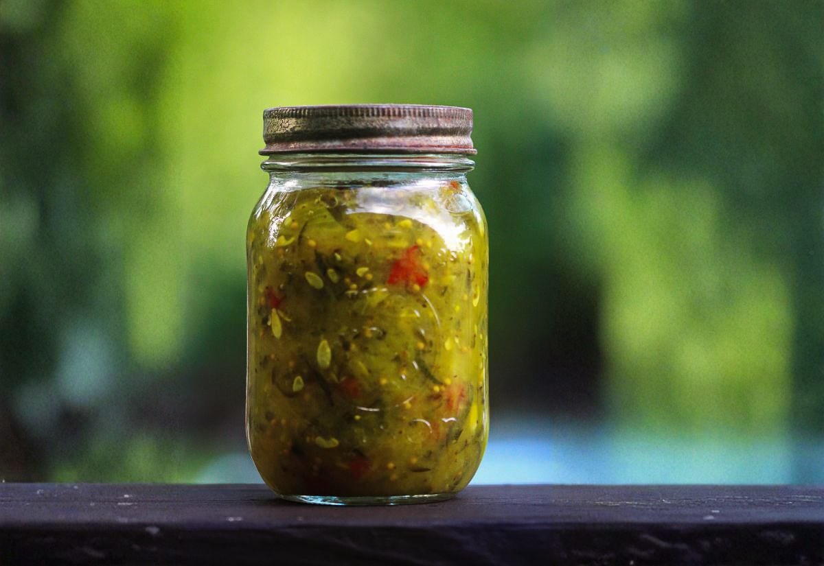 A glass mason jar filled with homemade green tomato relish, flecked with bits of red pepper, sitting on a dark wooden railing with a blurred garden background.
