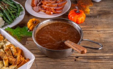 Small pot of dark, pan-dripping gravy on a rustic wooden table, surrounded by Thanksgiving sides like sliced ham, green beans, stuffing, and fall decorations.