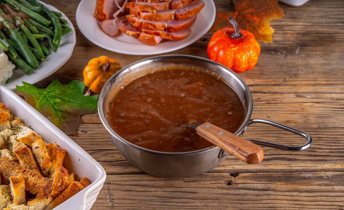 Small pot of dark, pan-dripping gravy on a rustic wooden table, surrounded by Thanksgiving sides like sliced ham, green beans, stuffing, and fall decorations.