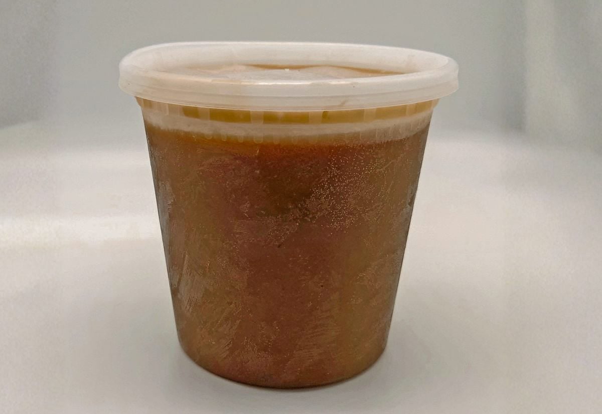 Frozen homemade gravy stored in a clear deli container, showing the solidified brown gravy and separated fat layer.