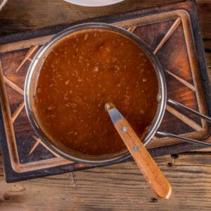 Overhead view of a pot filled with smooth, dark pan-dripping gravy resting on a rustic wooden board.