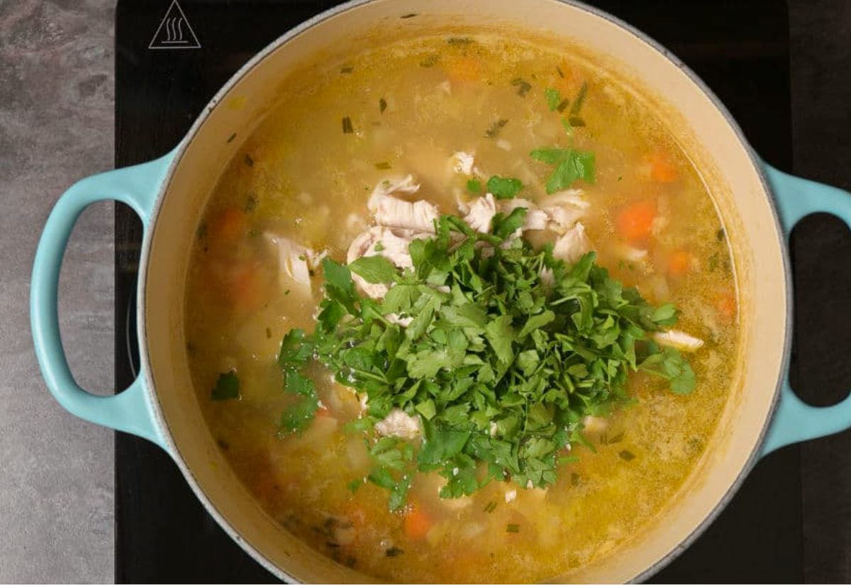 A large blue Dutch oven filled with chicken and barley soup, topped with a handful of fresh parsley. Chunks of chicken, carrots, and barley are visible in the golden broth, ready to stir and serve.