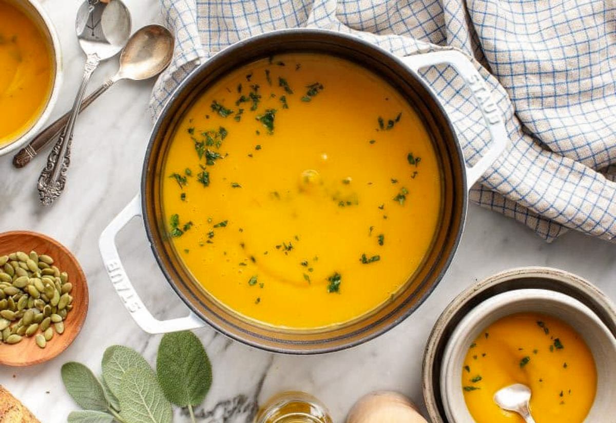 A pot of creamy butternut squash soup garnished with chopped herbs. The golden-orange soup sits on a marble counter surrounded by fresh sage leaves, pepitas, and a kitchen towel, ready to serve.