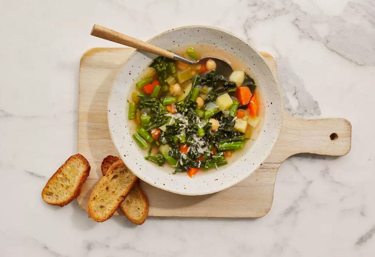 A bowl of colorful vegetable soup filled with kale, carrots, green beans, potatoes, and chickpeas, set on a wooden board with toasted bread on the side. The light broth and vibrant vegetables show its wholesome, homemade style.