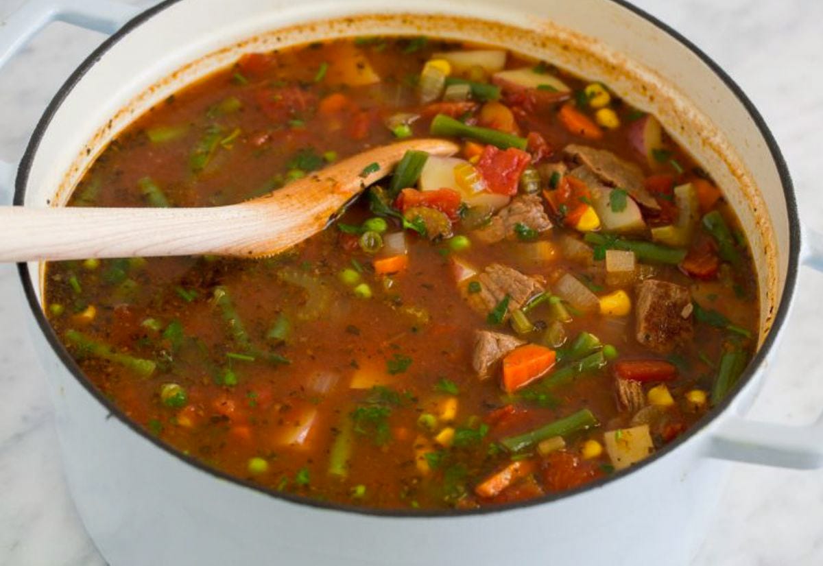 A Dutch oven filled with hearty vegetable beef soup made with tender chunks of beef, green beans, corn, carrots, and tomatoes in a rich red broth. A wooden spoon rests in the pot, ready to serve.