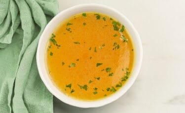 A white bowl filled with golden homemade turkey broth sprinkled with chopped parsley, sitting beside a soft green cloth on a light countertop — showing off the clear, rich color of well-skimmed stock.