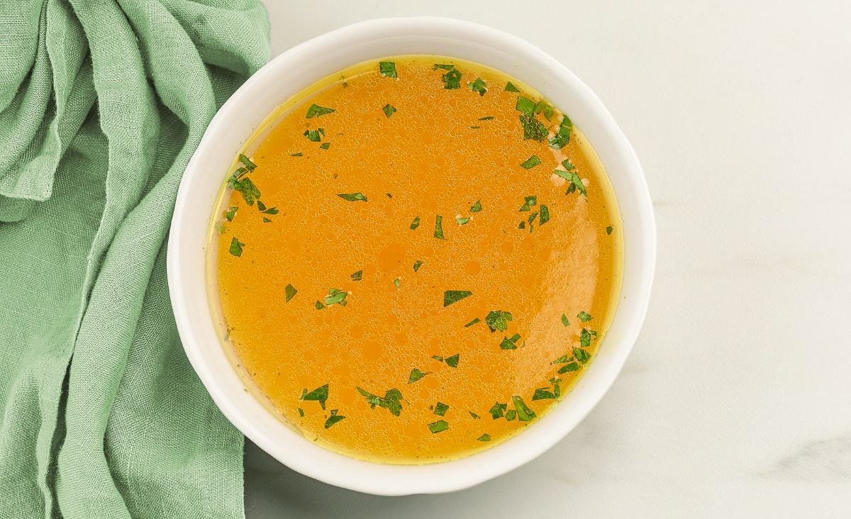 A white bowl filled with golden homemade turkey broth sprinkled with chopped parsley, sitting beside a soft green cloth on a light countertop — showing off the clear, rich color of well-skimmed stock.