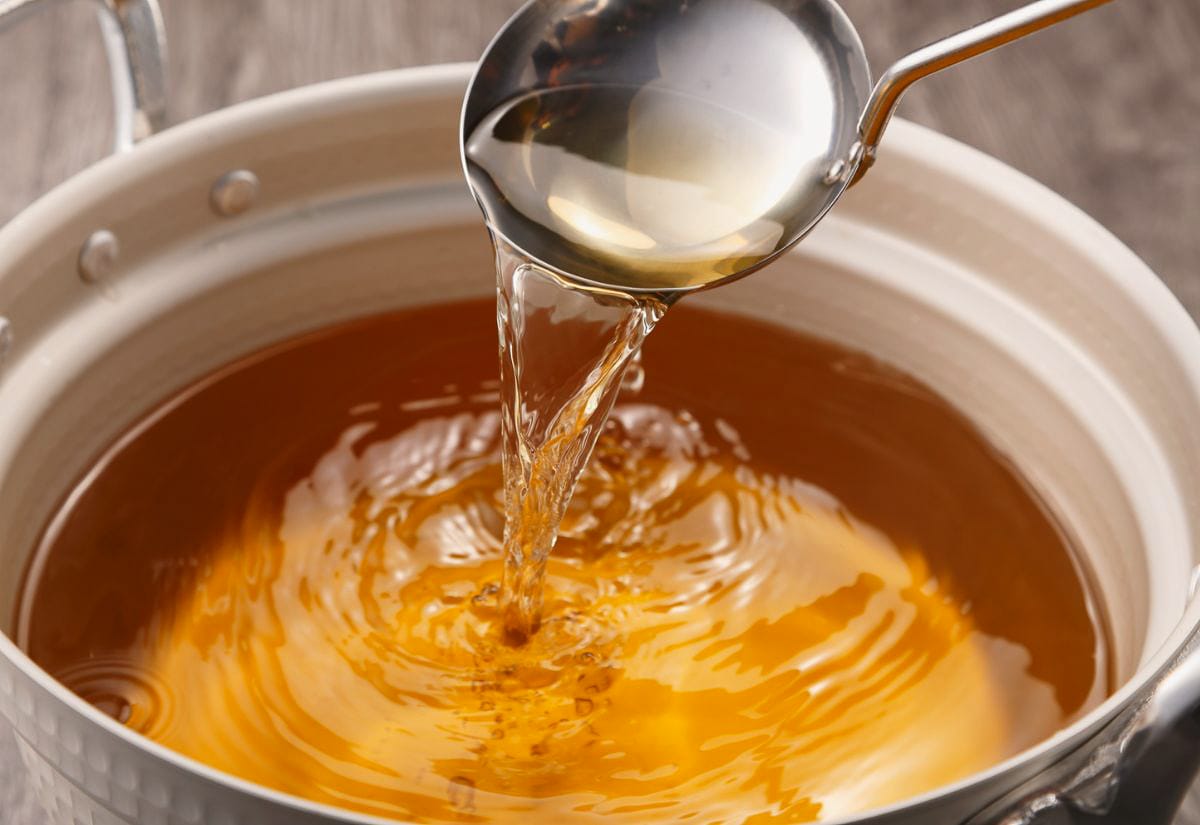 A ladle pouring golden, clear turkey stock back into a white enamel pot, catching warm light that highlights the broth’s clarity and smooth surface.