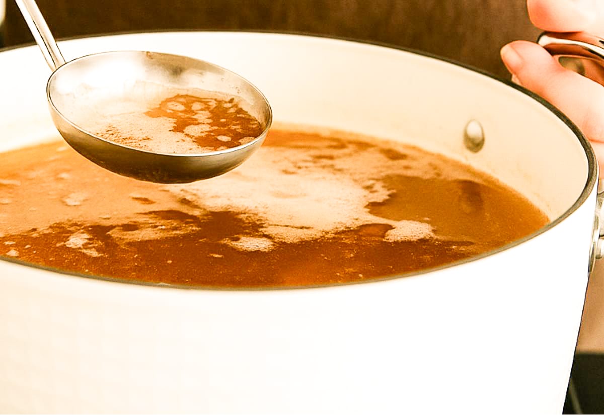 A close-up of a person skimming foam from simmering turkey broth using a stainless-steel ladle, with gentle steam rising from the white stockpot — demonstrating the process for achieving a clear stock.