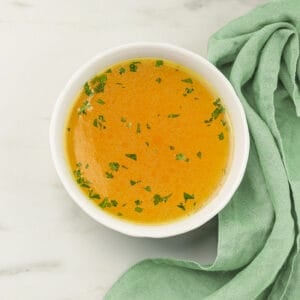 A white bowl of golden, clarified turkey broth garnished with herbs, viewed from above with a soft green linen napkin — the finished result of pressure-canning clear, shelf-stable stock.