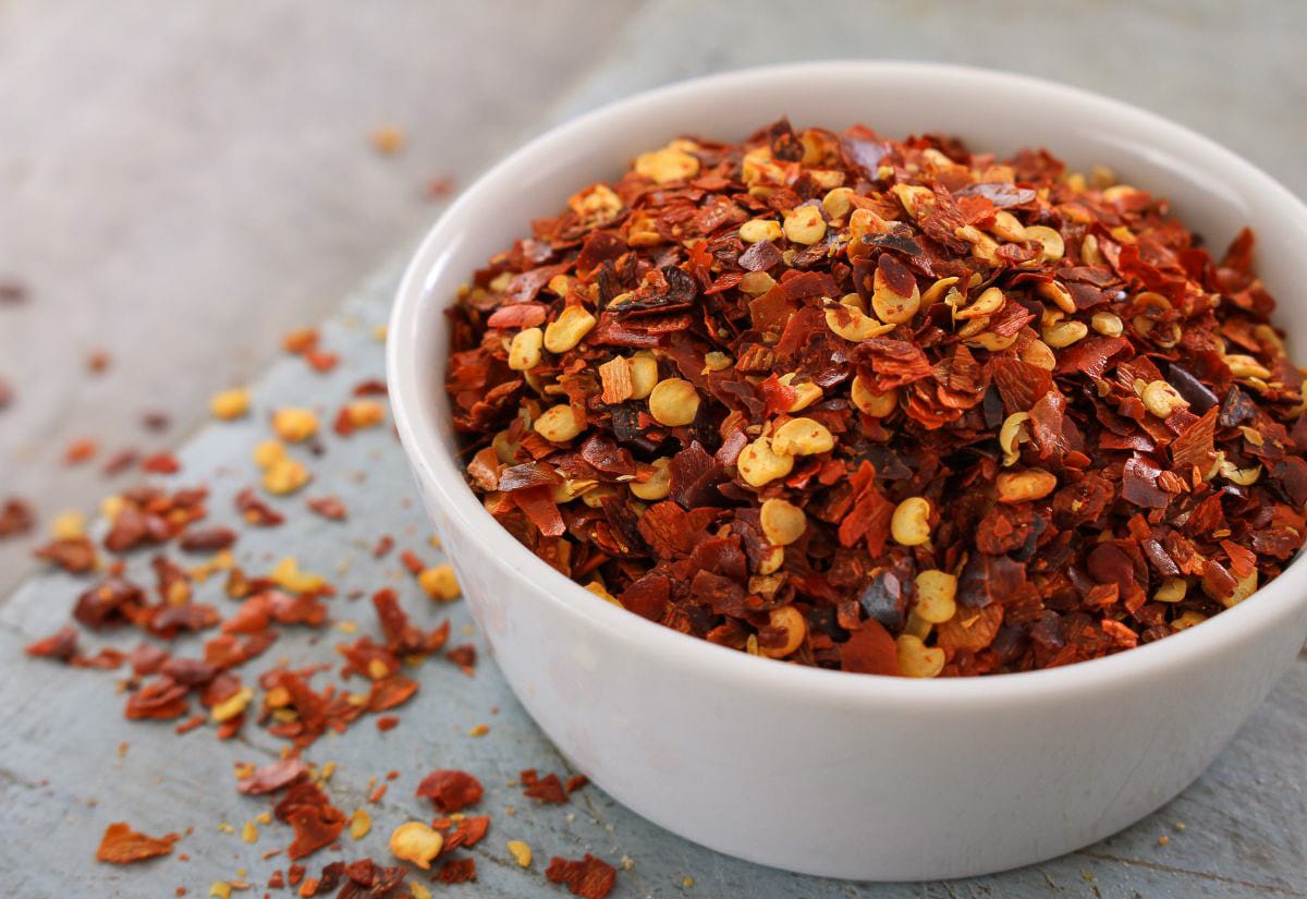 White bowl filled with red pepper flakes, with loose flakes scattered across a rustic surface, showing the texture and color of dried peppers.