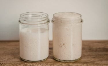 Two glass jars of sourdough starter sitting on a wooden counter; the jar on the left is less active with small bubbles, while the jar on the right is fuller and more bubbly, showing different stages of activity.