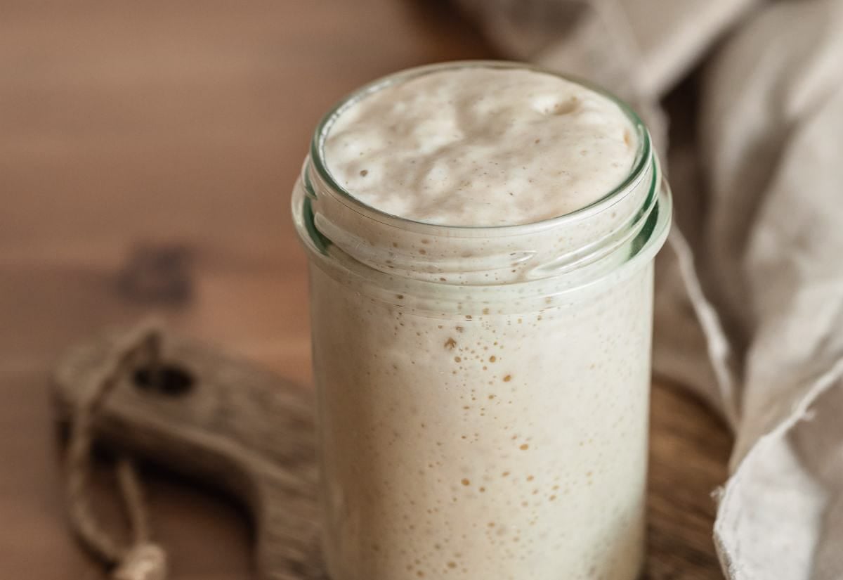 A single glass jar of freshly fed sourdough starter rising high in the jar, placed on a wooden cutting board with a neutral kitchen towel in the background.