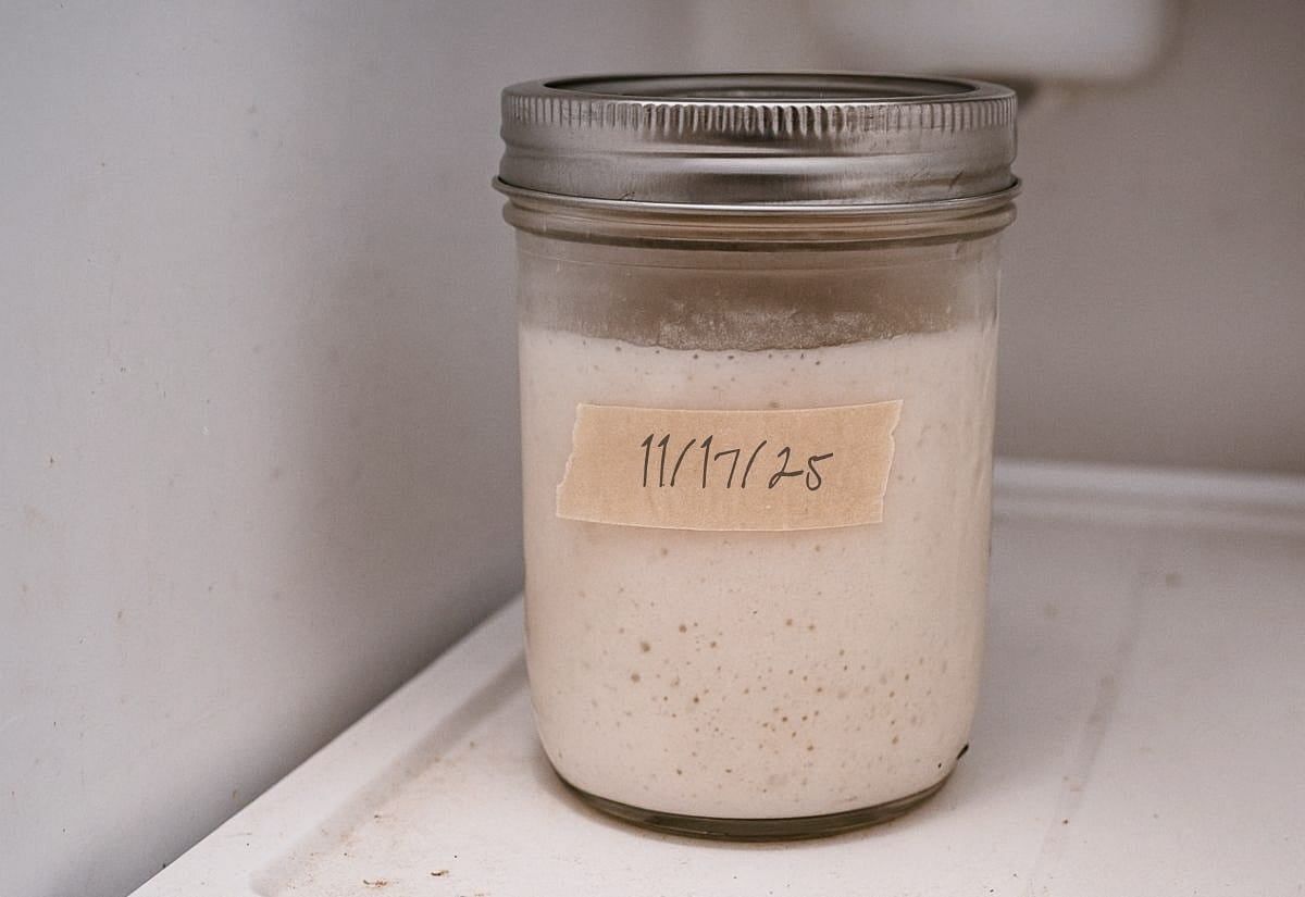 Glass jar of sourdough starter stored in the refrigerator, sealed with a metal lid and labeled “11/17/25” on a piece of masking tape, showing a moderately active starter.