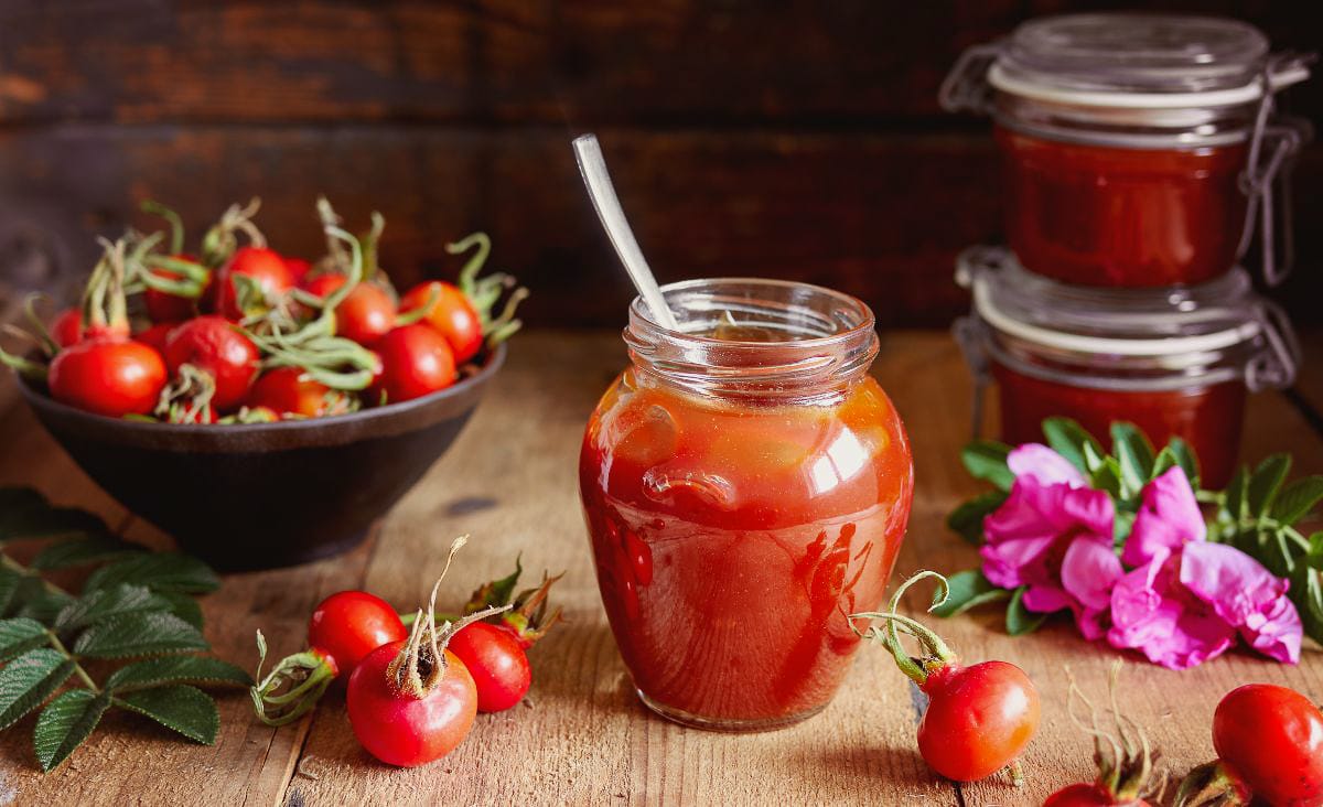 Close-up of a glass jar filled with bright orange rose hip jelly on a wooden table, surrounded by fresh rose hips, a bowl of fruit, and a pink rugosa rose blossom, capturing the cozy look of homemade foraged preserves.