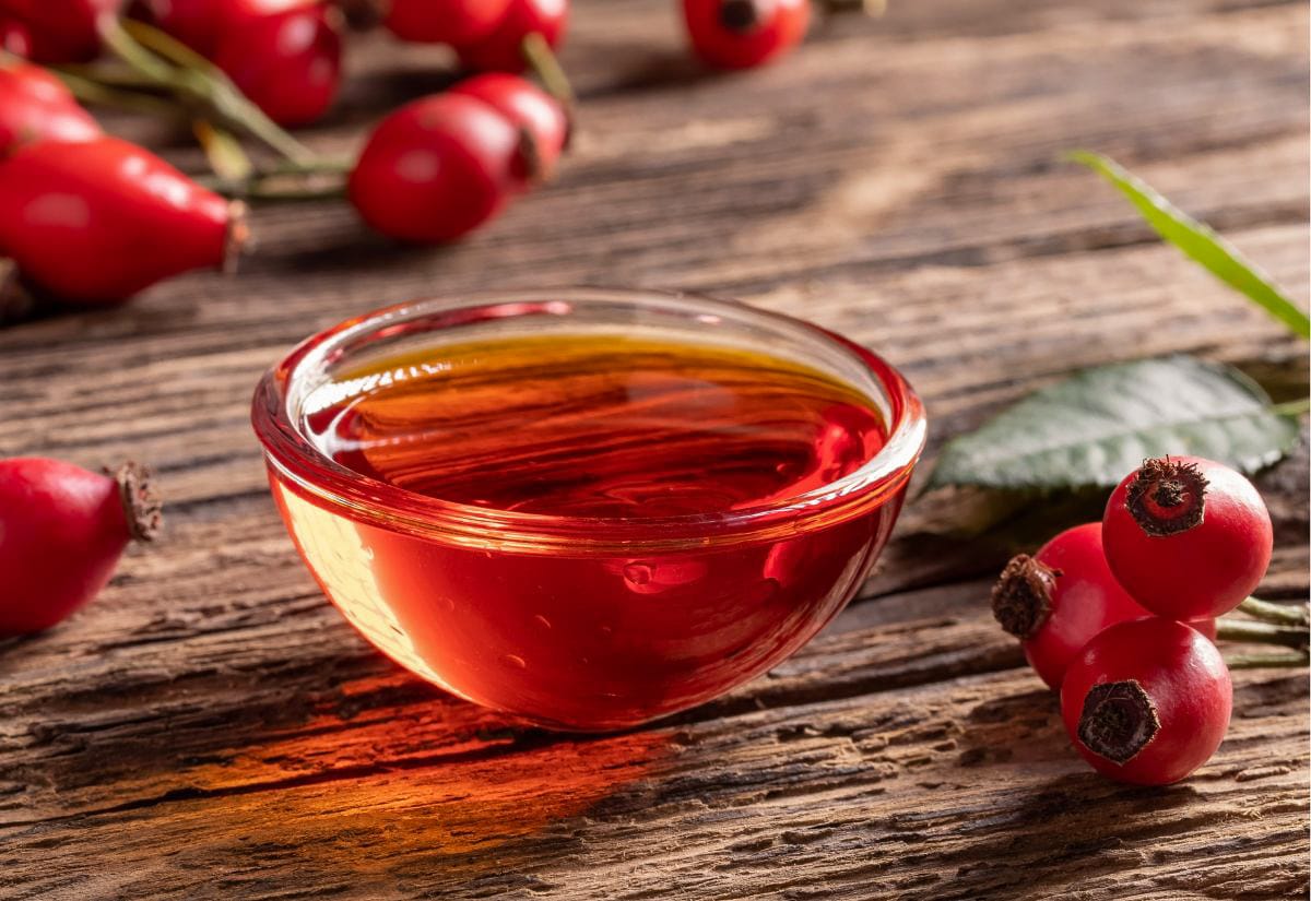 Glass bowl of translucent rose hip juice glowing amber in natural light on a wooden table, with fresh hips scattered nearby — showing the rich color before the jelly is made.