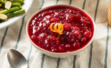 A small bowl filled with glossy cranberry relish garnished with orange peel, sitting next to a spoon and a plate of green beans on a striped kitchen towel.