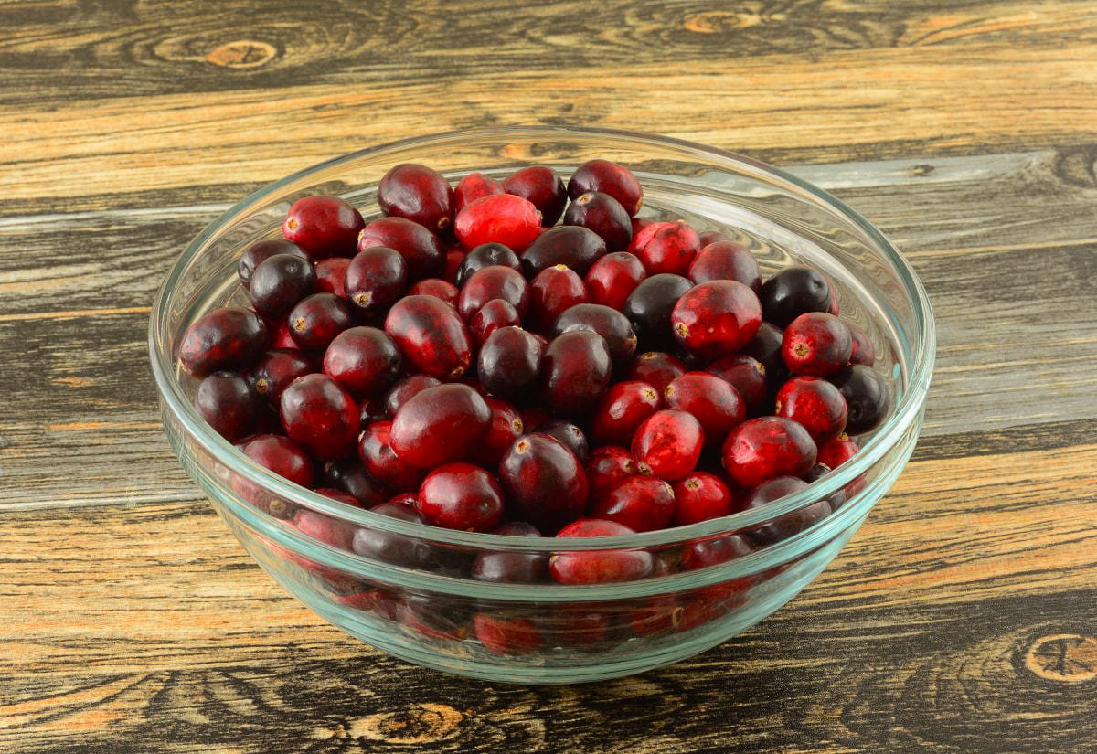 Clear glass bowl filled to the top with fresh whole cranberries, set on a rustic wood-grain surface.