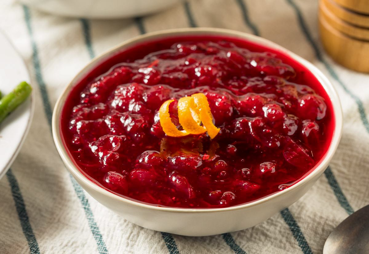 Close-up of a bowl of chunky cranberry relish with visible whole berries and an orange zest twist, sitting on a cream and blue striped cloth.