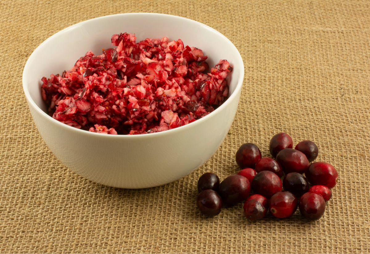 White ceramic bowl filled with freshly chopped cranberries for relish, with a small pile of whole cranberries placed beside it on burlap.
