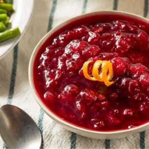 Close-up view of thick, glossy cranberry sauce topped with a twist of orange peel, served in a white bowl with vegetables slightly visible in the background.