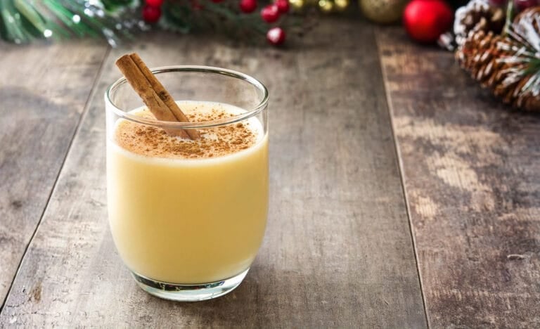 A glass of creamy homemade eggnog topped with cinnamon and garnished with a cinnamon stick, sitting on a weathered wooden table with soft holiday greenery blurred in the background.