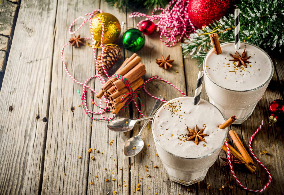 Two frothy holiday eggnog drinks topped with star anise and cinnamon sticks on a rustic wooden table surrounded by Christmas ornaments, pinecones, and twine.