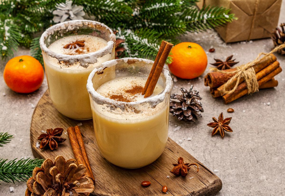Two glasses of homemade eggnog with sugar-frosted rims, cinnamon sticks, and a dusting of nutmeg, styled with fresh clementines, pine branches, and pinecones for a cozy holiday look.