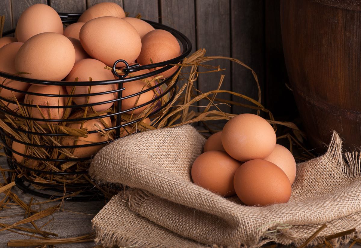 A wire basket overflowing with fresh brown eggs resting in straw, with a small pile of eggs set on burlap beside it in a rustic homestead kitchen scene.