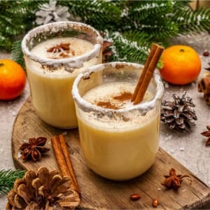 Two glasses of creamy homemade eggnog with frosted rims and cinnamon sticks, set on a small wooden board surrounded by pinecones, star anise, and bright clementines.