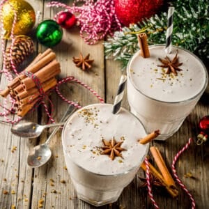Two thick eggnog drinks topped with star anise and served with striped straws, surrounded by cinnamon sticks, ornaments, and evergreen branches on a rustic wooden table.