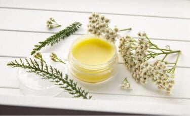 Small glass jar filled with pale yellow homemade hand salve on a white wooden surface, surrounded by fresh yarrow flowers and feathery yarrow leaves.