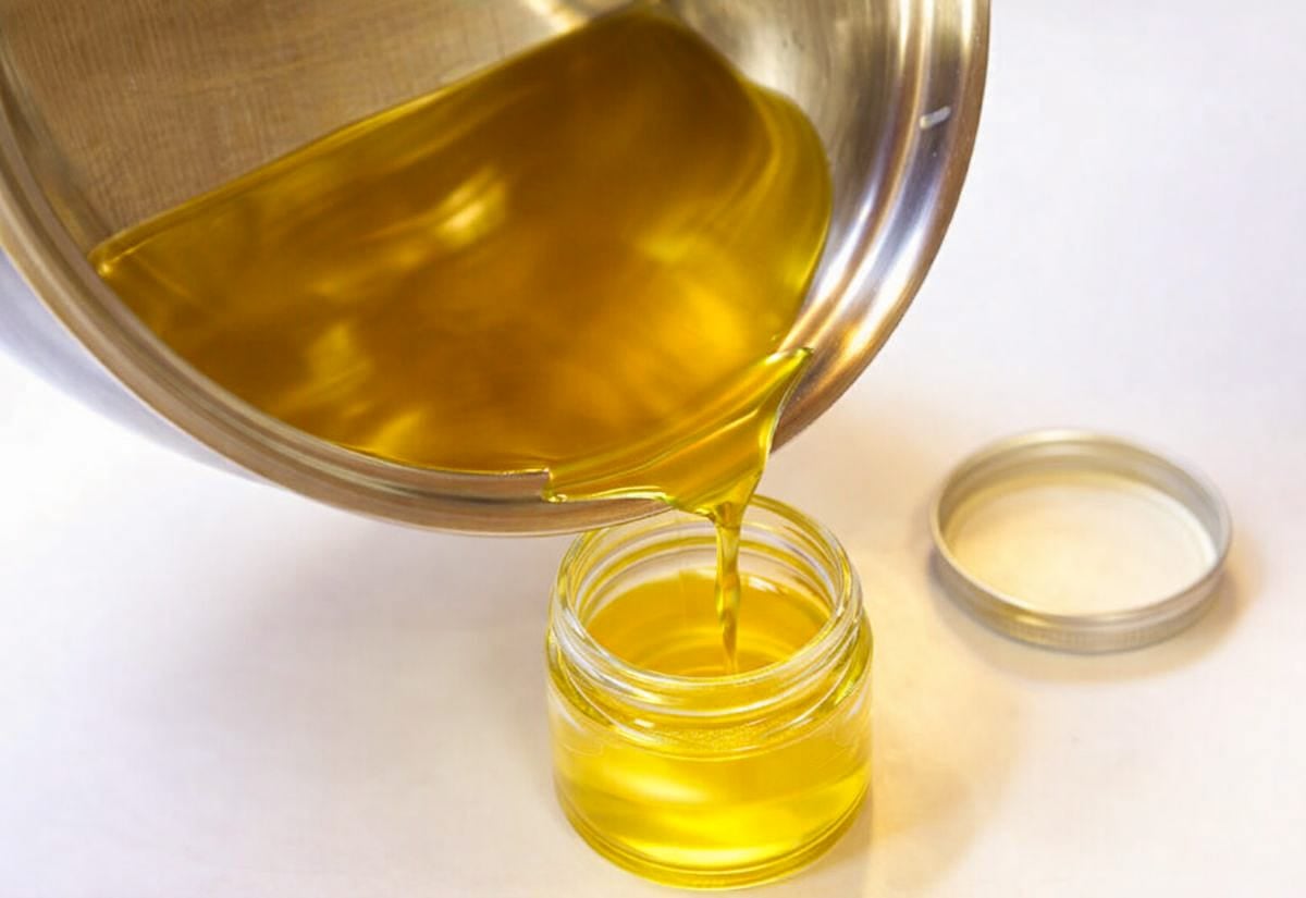 Warm homemade hand salve being poured from a stainless steel pot into a small glass jar, showing the golden-yellow color and liquid texture before it sets.