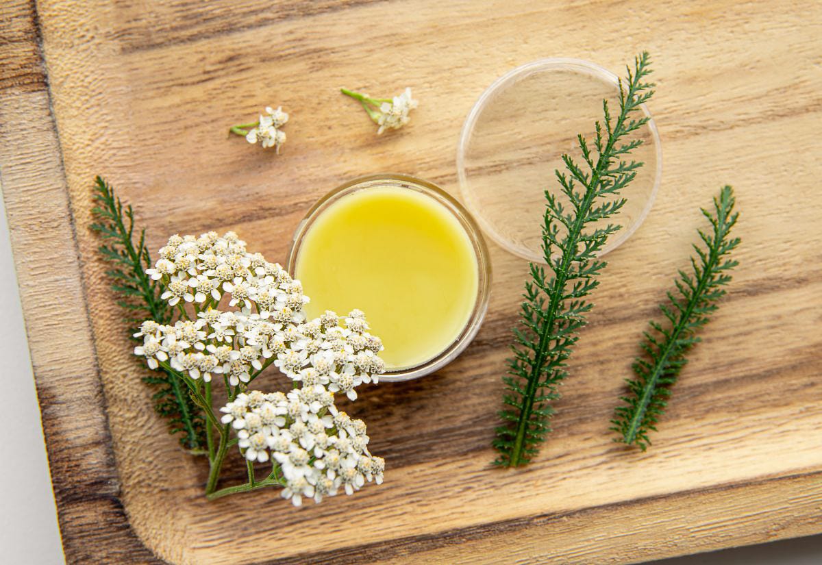 Top-down view of a jar of homemade yarrow hand salve on a wooden cutting board, surrounded by fresh yarrow flowers and green yarrow leaves.