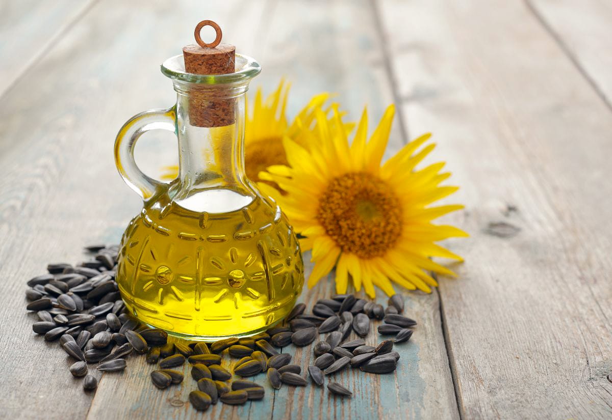 Glass bottle of sunflower oil surrounded by sunflower seeds and bright yellow sunflower heads, representing the lightweight oil used in homemade hand salve.