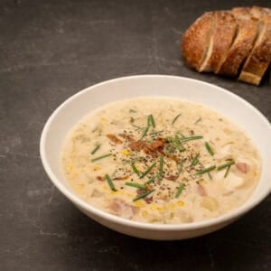 A simple bowl of Maine-style corn chowder with visible potatoes and corn, photographed with plenty of negative space and bread in the background.