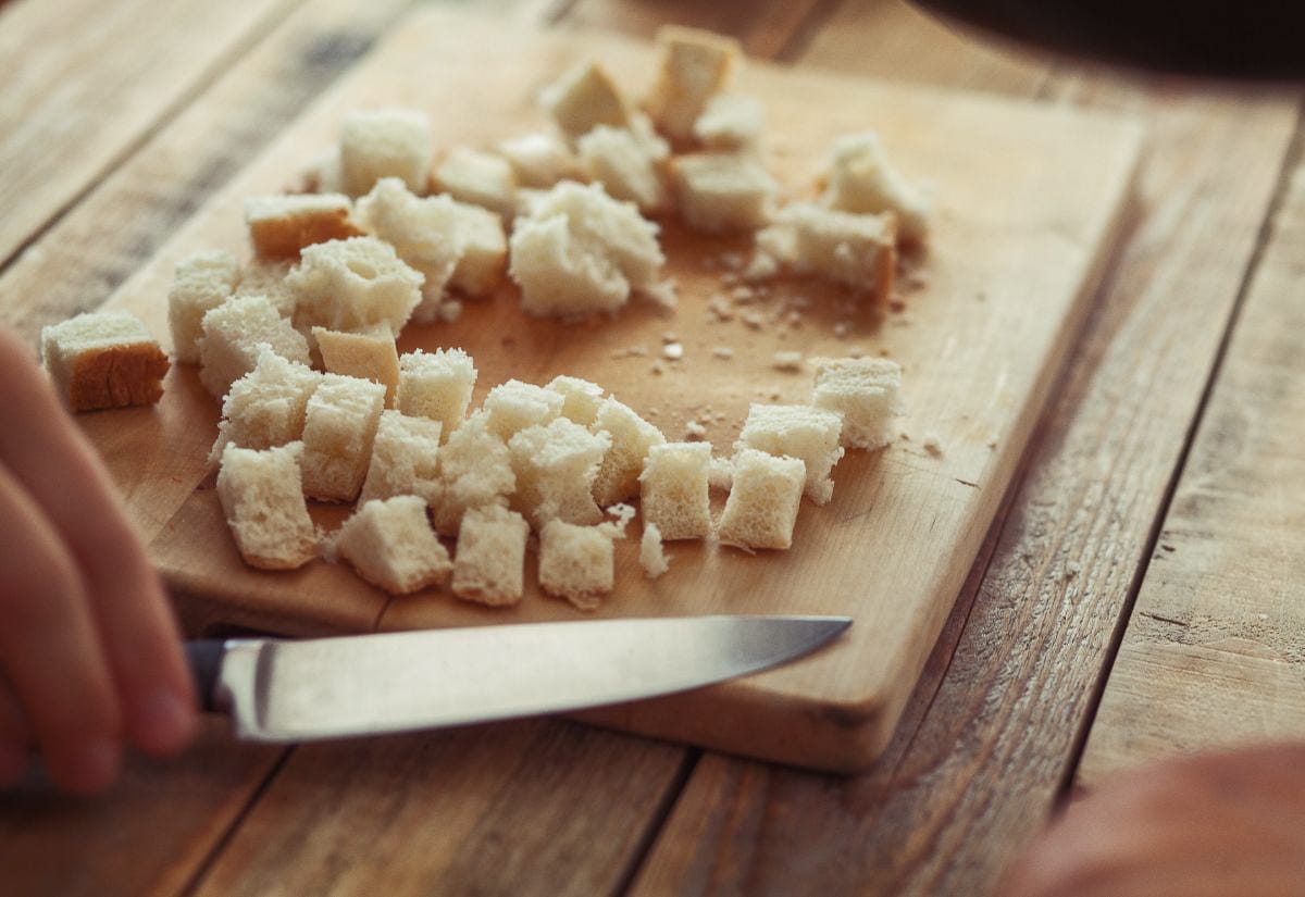Small bread cubes being cut on a rustic wooden cutting board with a knife, showing the preparation step before baking homemade croutons.