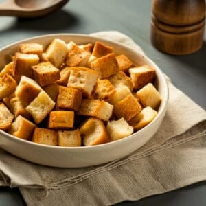 A bowl filled with golden, crispy croutons made from stale bread, set on a folded kitchen towel with warm lighting.