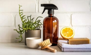 Amber glass spray bottle, lemon half, bar soap, scrub brush, folded cloths, and a small potted plant arranged on a light kitchen counter against a white brick wall.
