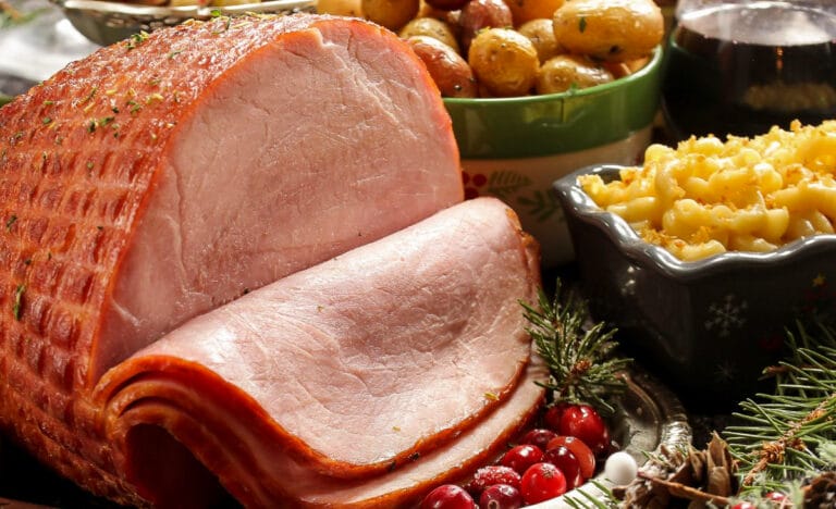 Glazed ham sliced on a serving platter surrounded by classic side dishes like roasted potatoes and macaroni and cheese.
