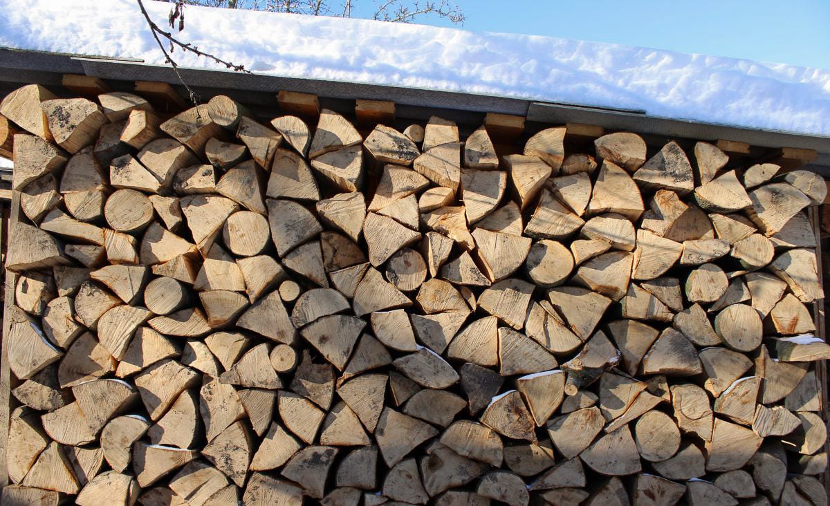 Wide outdoor woodpile stacked tightly under a simple wood shed roof covered in snow. The split logs are cut to uniform length and arranged bark-side out, forming a dense wall of seasoned firewood ready for winter heating.
