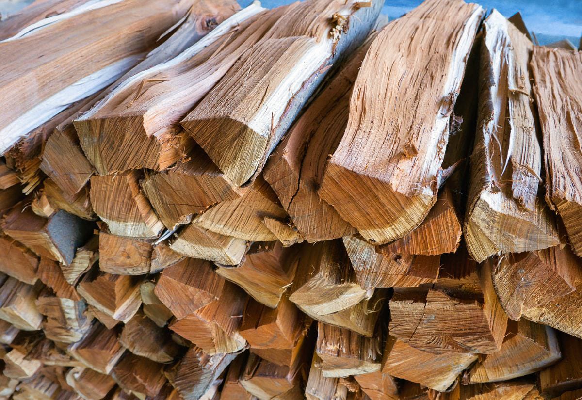 Close-up view of freshly split hardwood logs stacked in a crisscross pattern, showing detailed wood grain, rough split edges, and varying shades of tan and golden brown. The texture and tight stacking highlight properly prepared firewood for home heating.