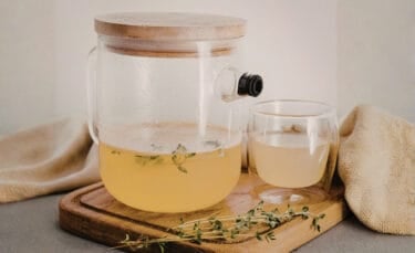 Glass pitcher with built-in infuser steeping light-colored herbal tea, fresh herb sprigs inside, sitting on a wooden board next to a double-walled glass cup and beige cloth.