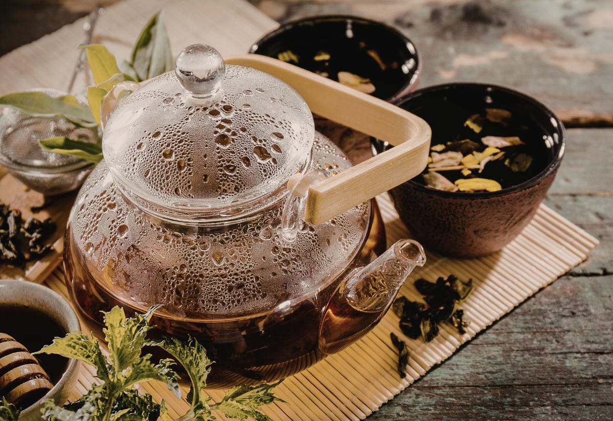 Clear glass teapot filled with hot herbal tea and condensation on the lid, surrounded by loose dried herbs, small bowls, and a honey dipper on a rustic surface.